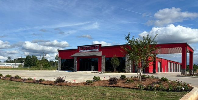 Red commercial building with a black awning and sign, set beside a parking lot and landscaped grass under a blue sky