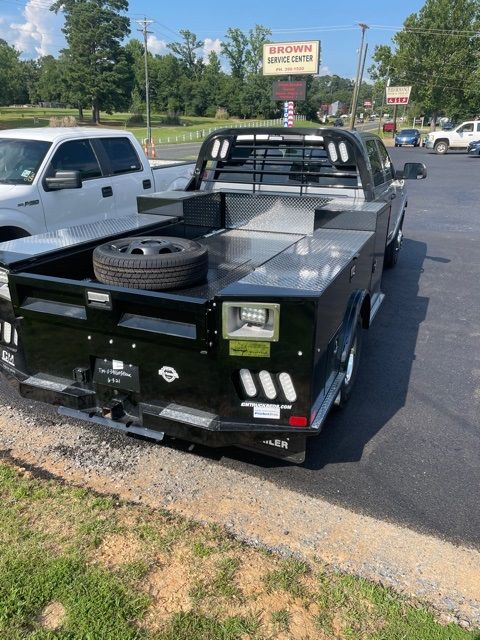 Black flatbed utility truck parked by the road, with a spare tire on the rear deck and a white pickup beside it.