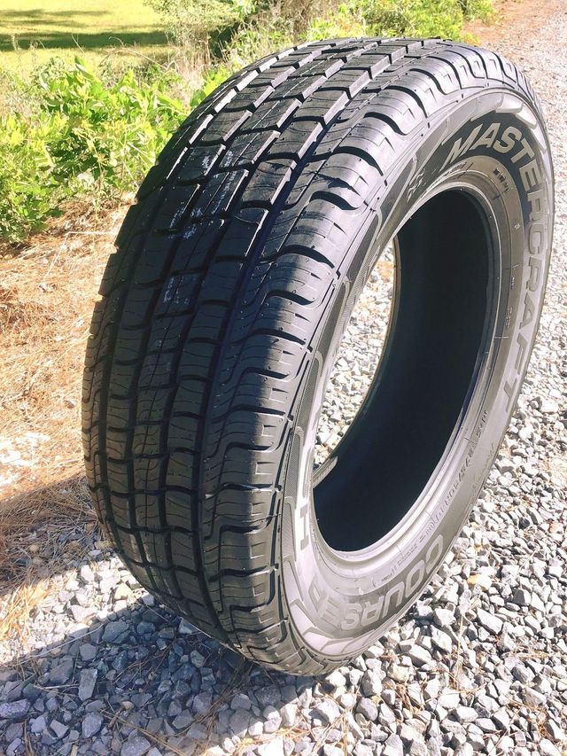 Large black tire standing upright on gravel outdoors in sunlight, with grass in the background