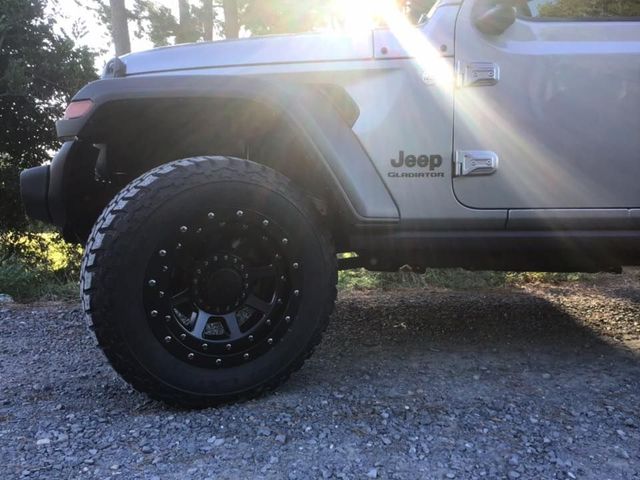 Silver Jeep Wrangler with black off-road wheel, parked on gravel in bright sunlight.