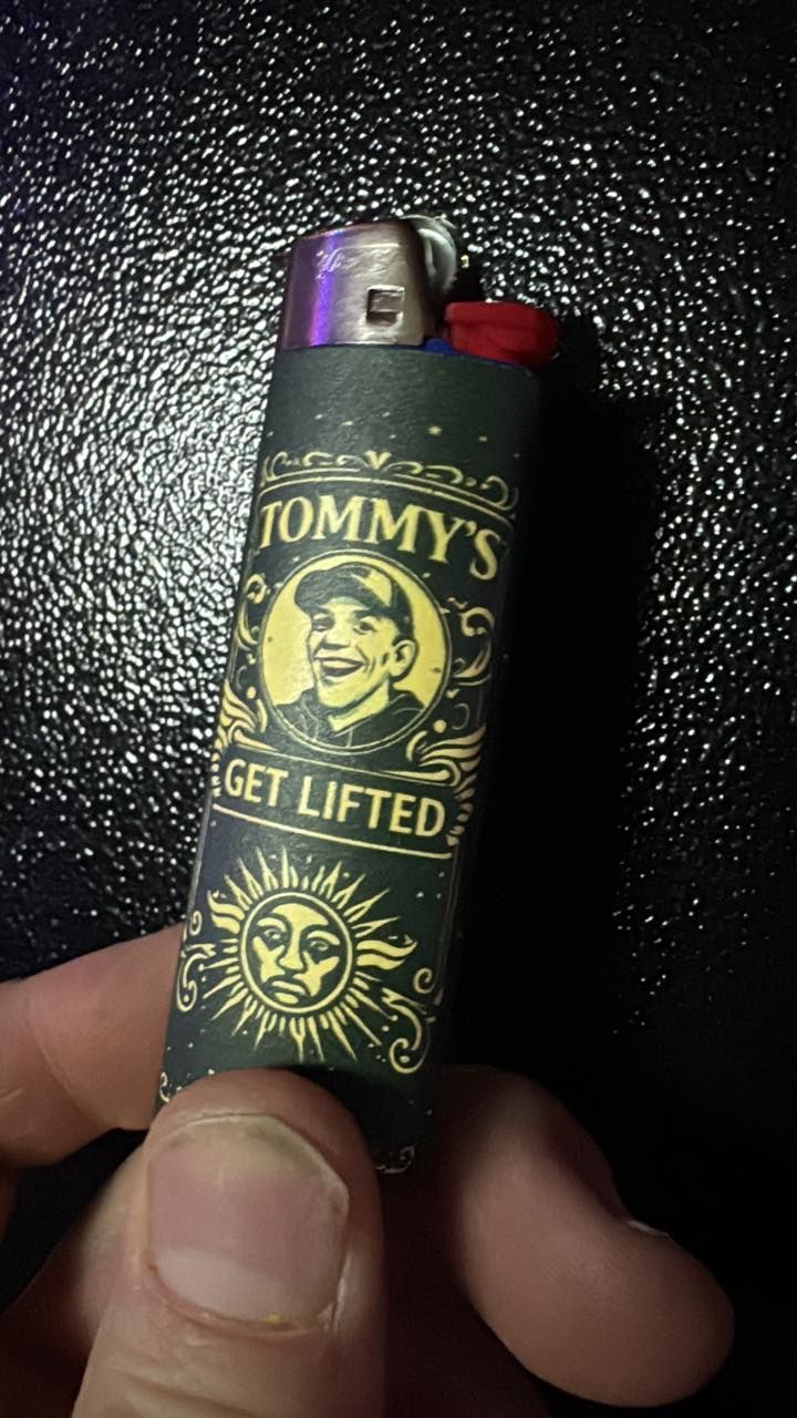 A hand holds a black and gold Tommy’s “Get Lifted” lighter against a dark background.
