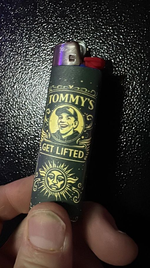 Hand holding a black and gold Tommy’s “Get Lifted” lighter against a dark background