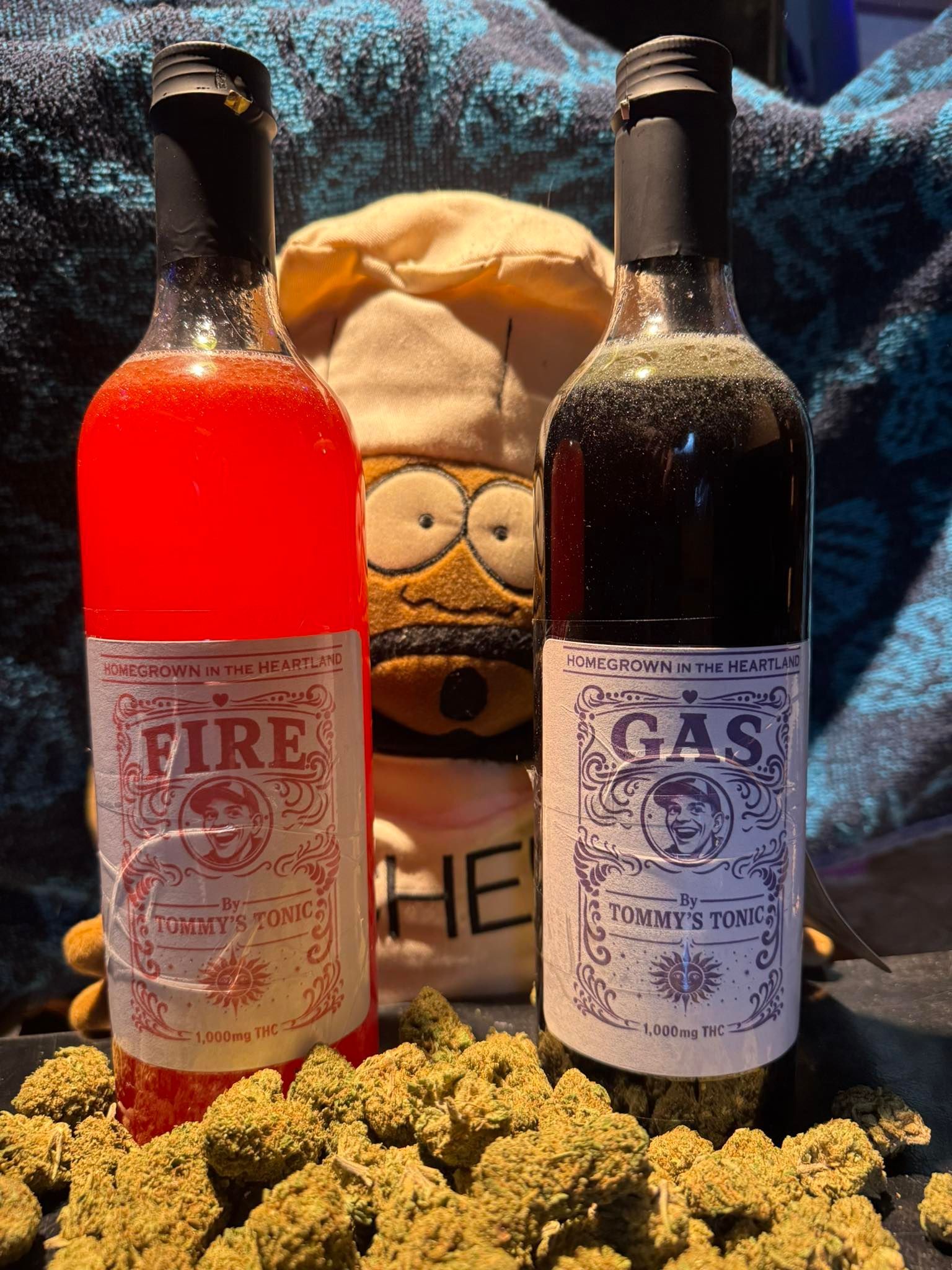 Two labeled bottles, “FIRE” and “GAS,” standing before a face-like plush and cannabis buds.