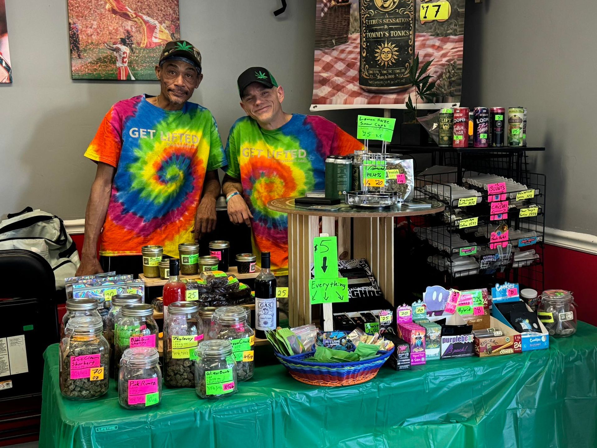 Two people behind a table of colorful packaged goods and jars at a market booth with tie-dye shirts