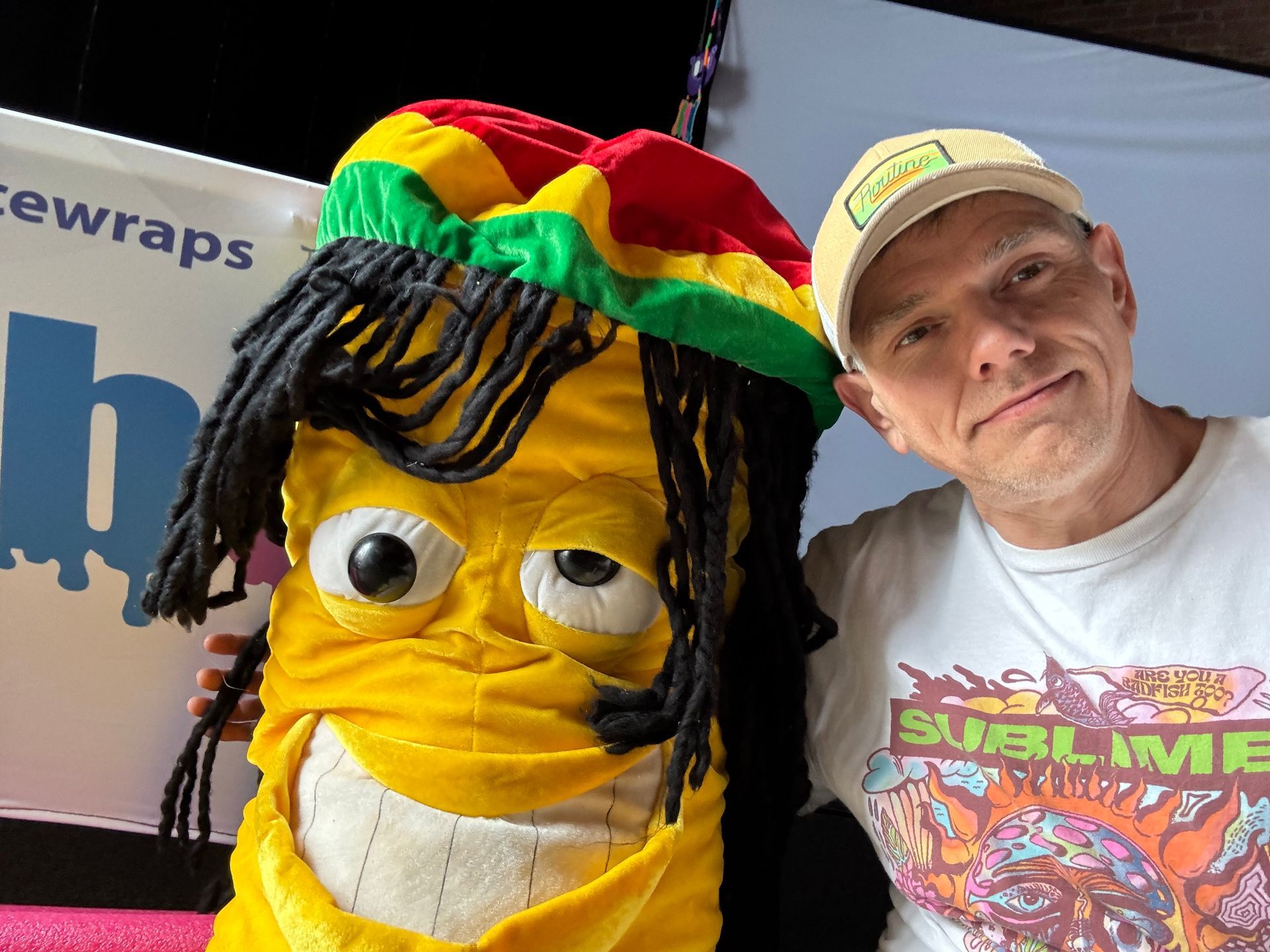 Smiling man beside a yellow puppet with dreadlocks and a red-green-yellow hat at an event backdrop