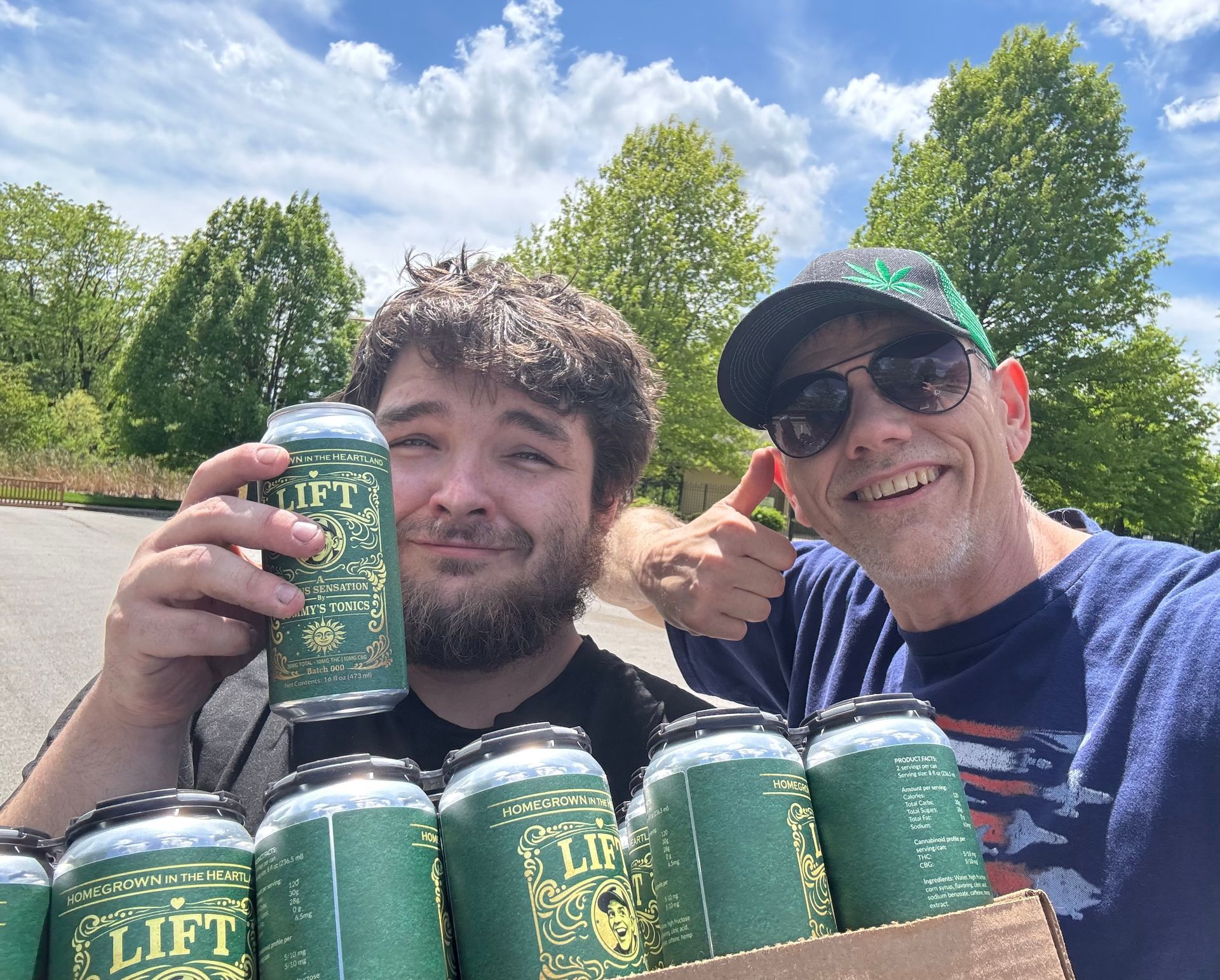 Two smiling people outdoors holding green cans, with a cardboard tray of cans in front of them.