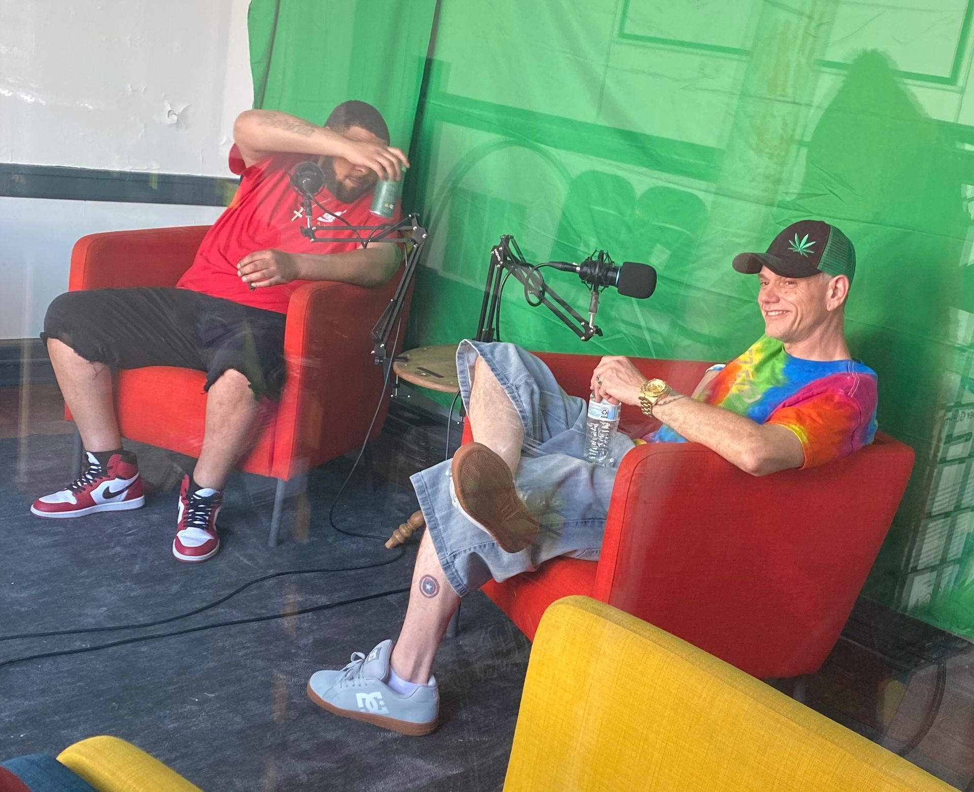 Two people lounging on red chairs with a microphone setup in front of a green backdrop.