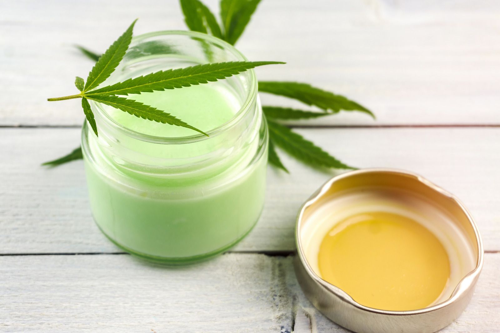 Green cream jar with cannabis leaf beside a small bowl of yellow oil on a white wooden surface