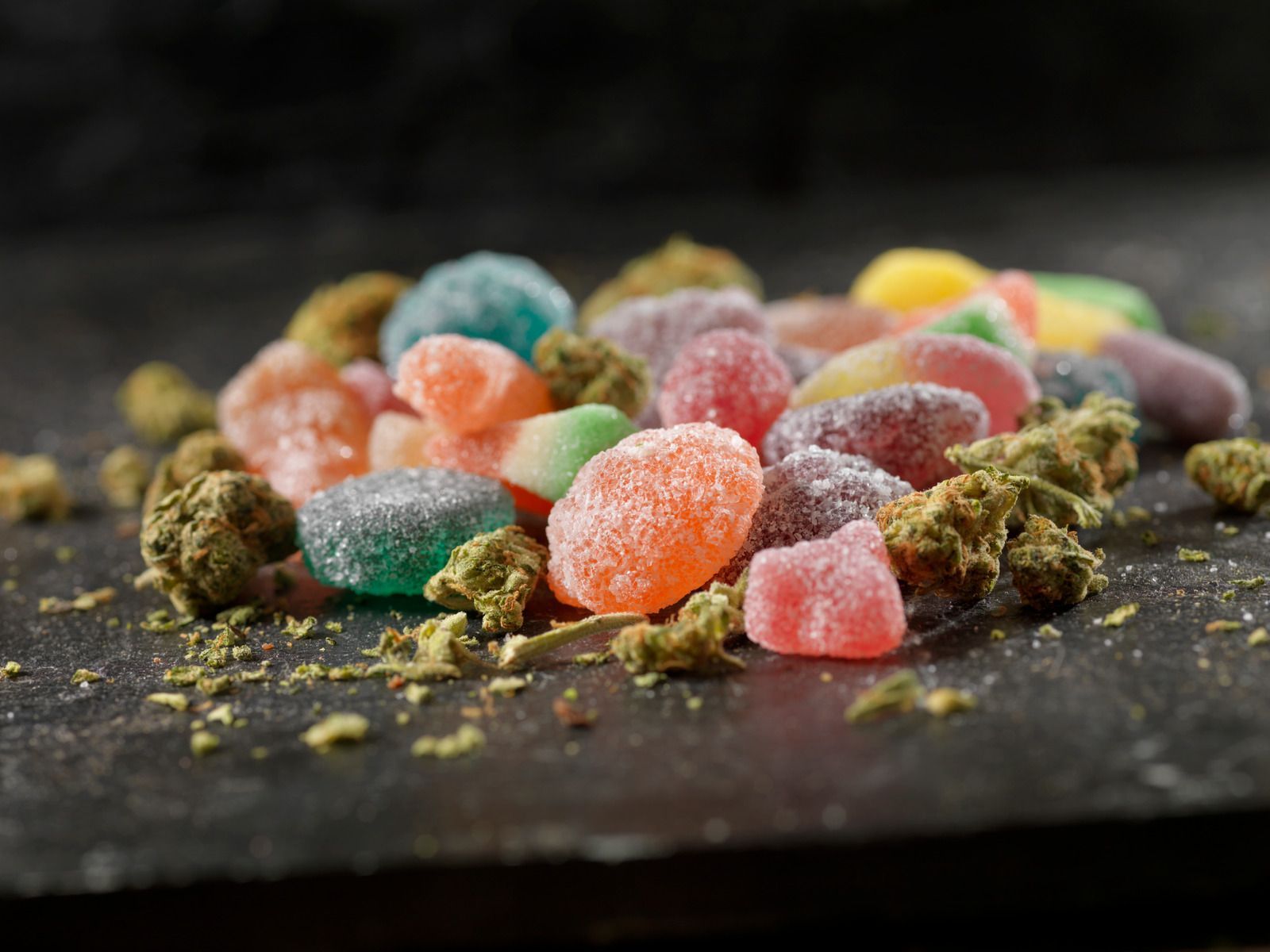 Colorful sugar-coated gummy candies scattered on a dark surface with cannabis buds nearby