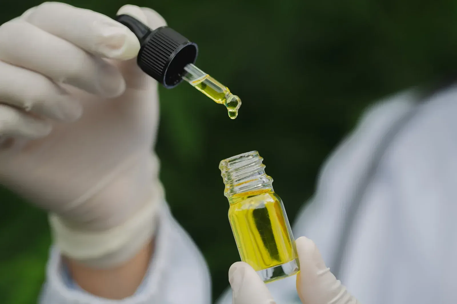 Gloved hands holding a dropper over a small bottle of yellow liquid in a lab setting