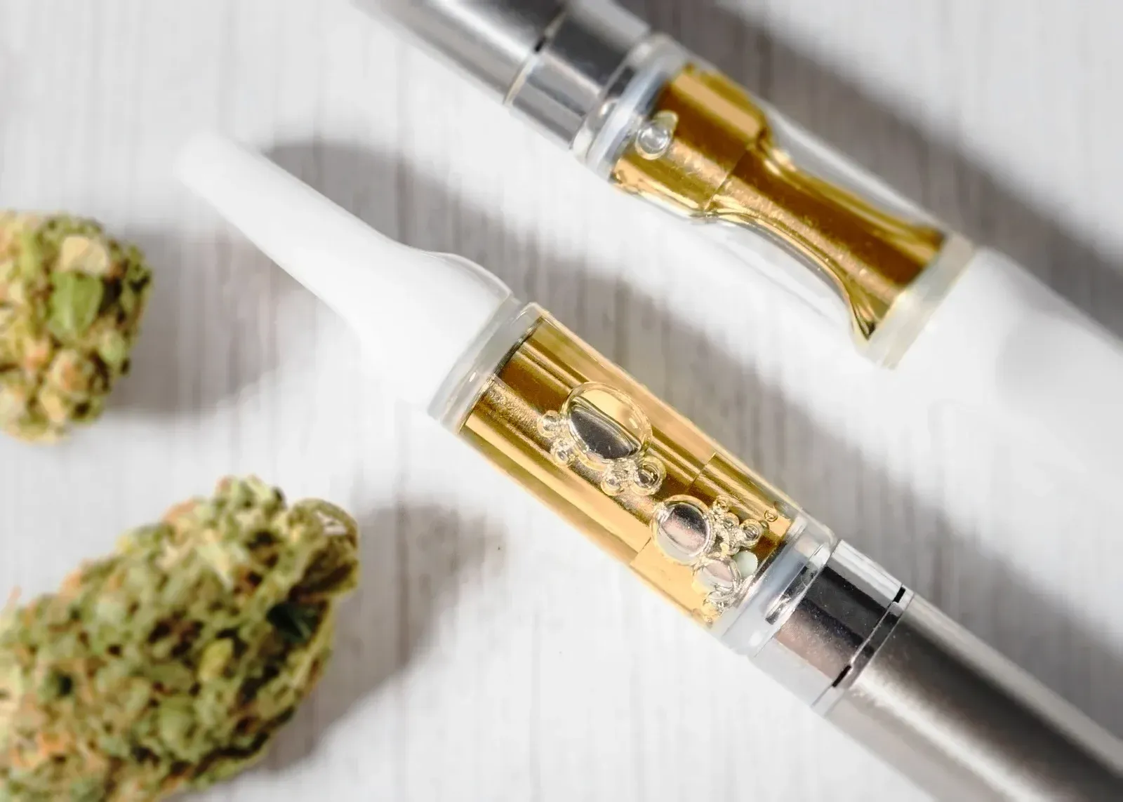 Two cannabis vape pens with clear oil cartridges beside cannabis buds