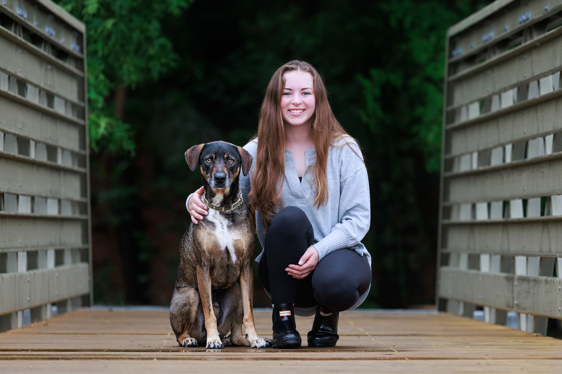 Young woman kneeling beside her dog on a wooden bridge during an outdoor pet photography session in 