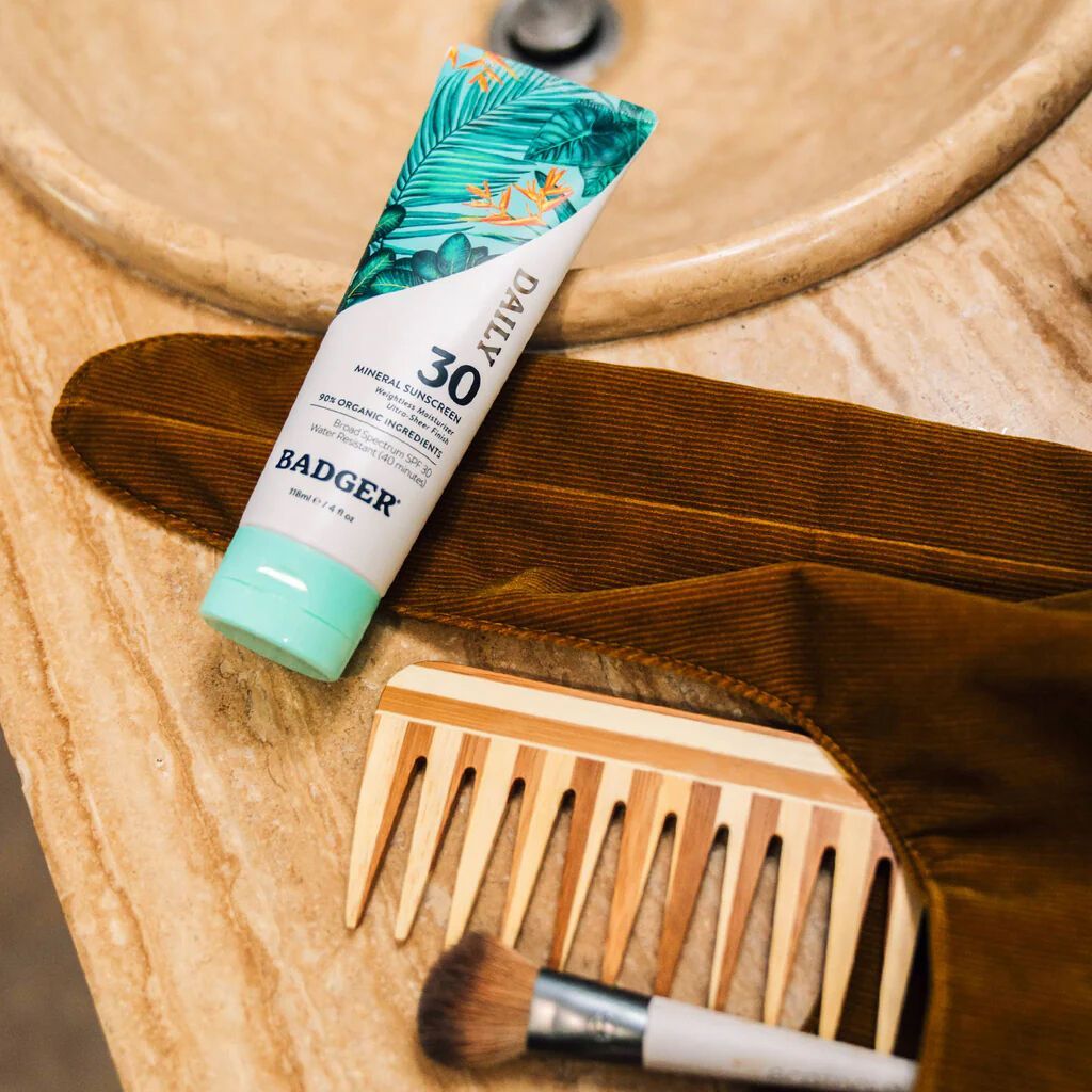 A bottle of badger sunscreen is sitting on a bathroom counter next to a comb and brush.