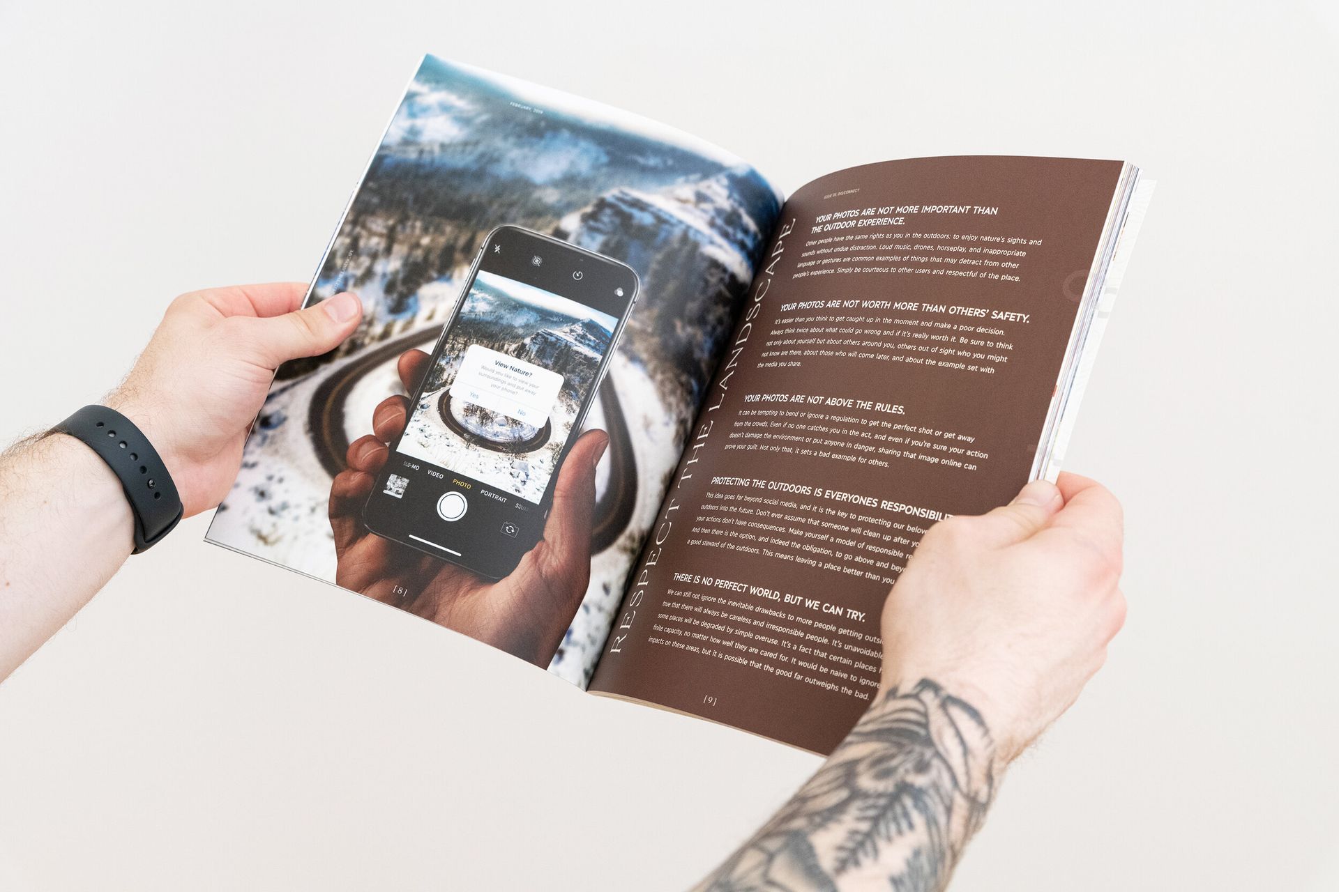 A person with a tattoo on their arm is holding a book and taking a picture of a cell phone.