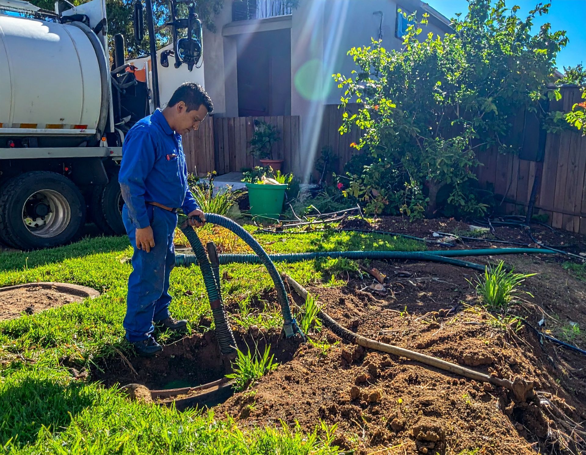 A worker in a blue uniform uses a large hose to pump a septic tank in a yard with a truck parked nearby.