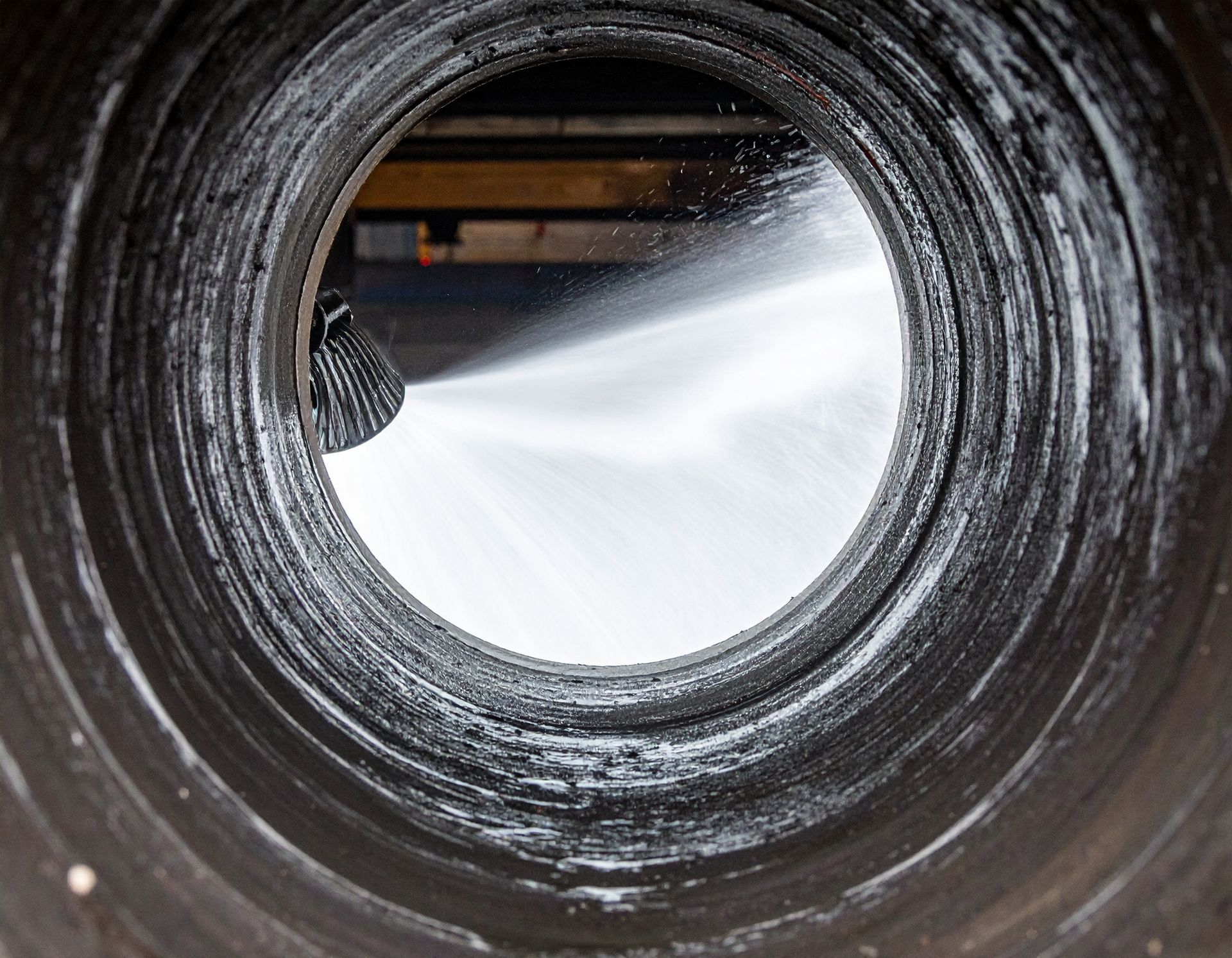 A close-up view through a dark, ridged pipe as a high-pressure water nozzle sprays a jet of water into the passage.