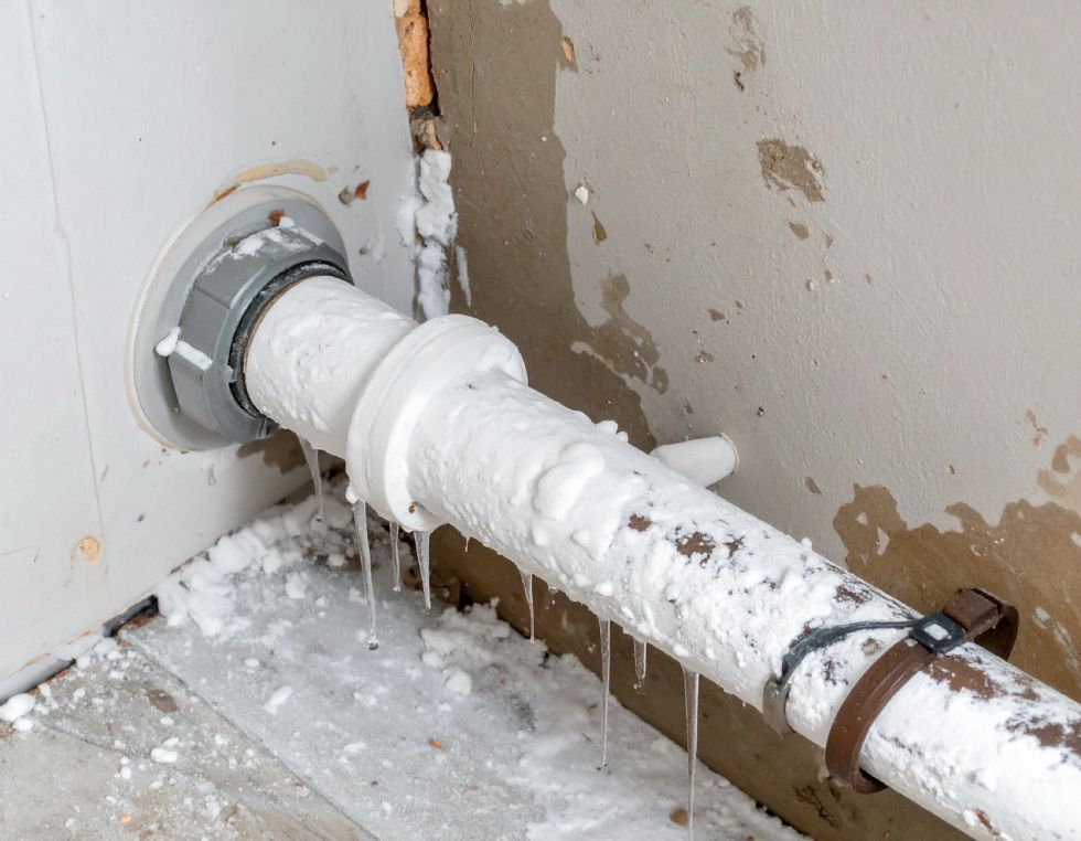 A white pipe protruding from a wall is covered in frost and icicles, indicating a frozen plumbing situation.