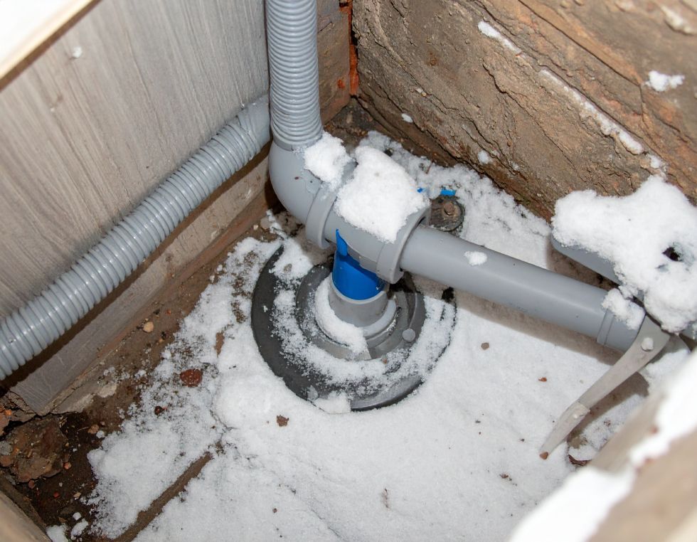 Grey pipes and a circular drain base are partially covered in snow against a wooden corner.