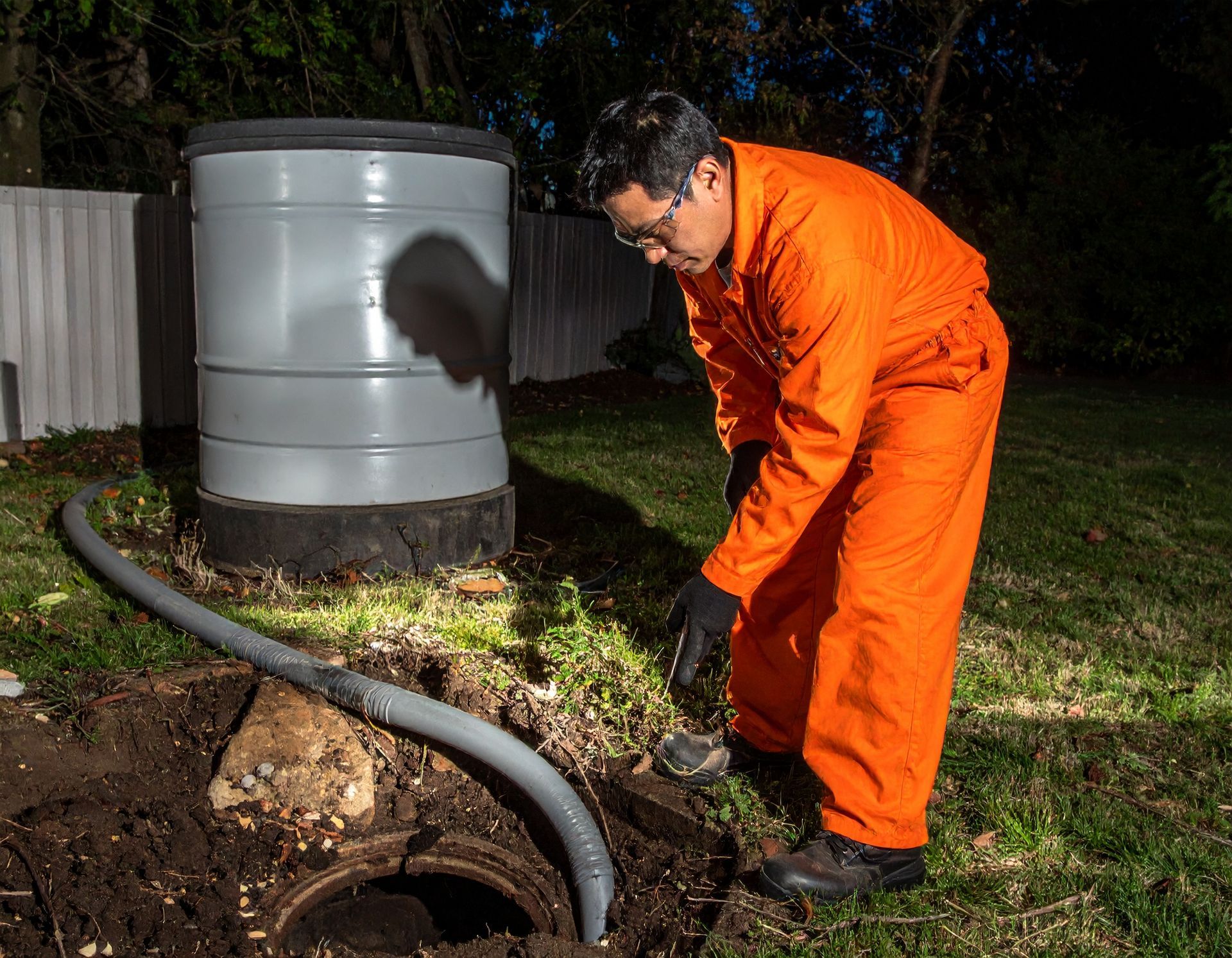 A person in an orange jumpsuit uses a hose to work on an open underground tank in a grassy yard.