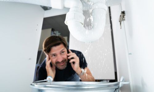 A person talks on a phone while looking distressed at water leaking from sink pipes into a metal bucket underneath.