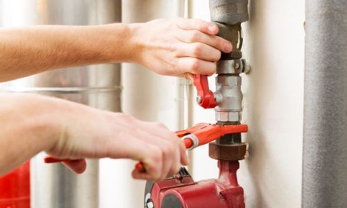Hands using red pliers to work on a metal pipe connection near a red pump in an industrial or basement setting.