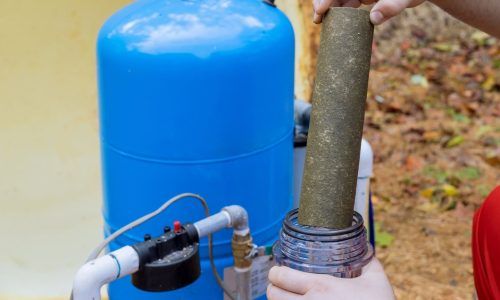 A person holds a dirty, sediment-covered water filter cartridge next to a blue residential well pressure tank.