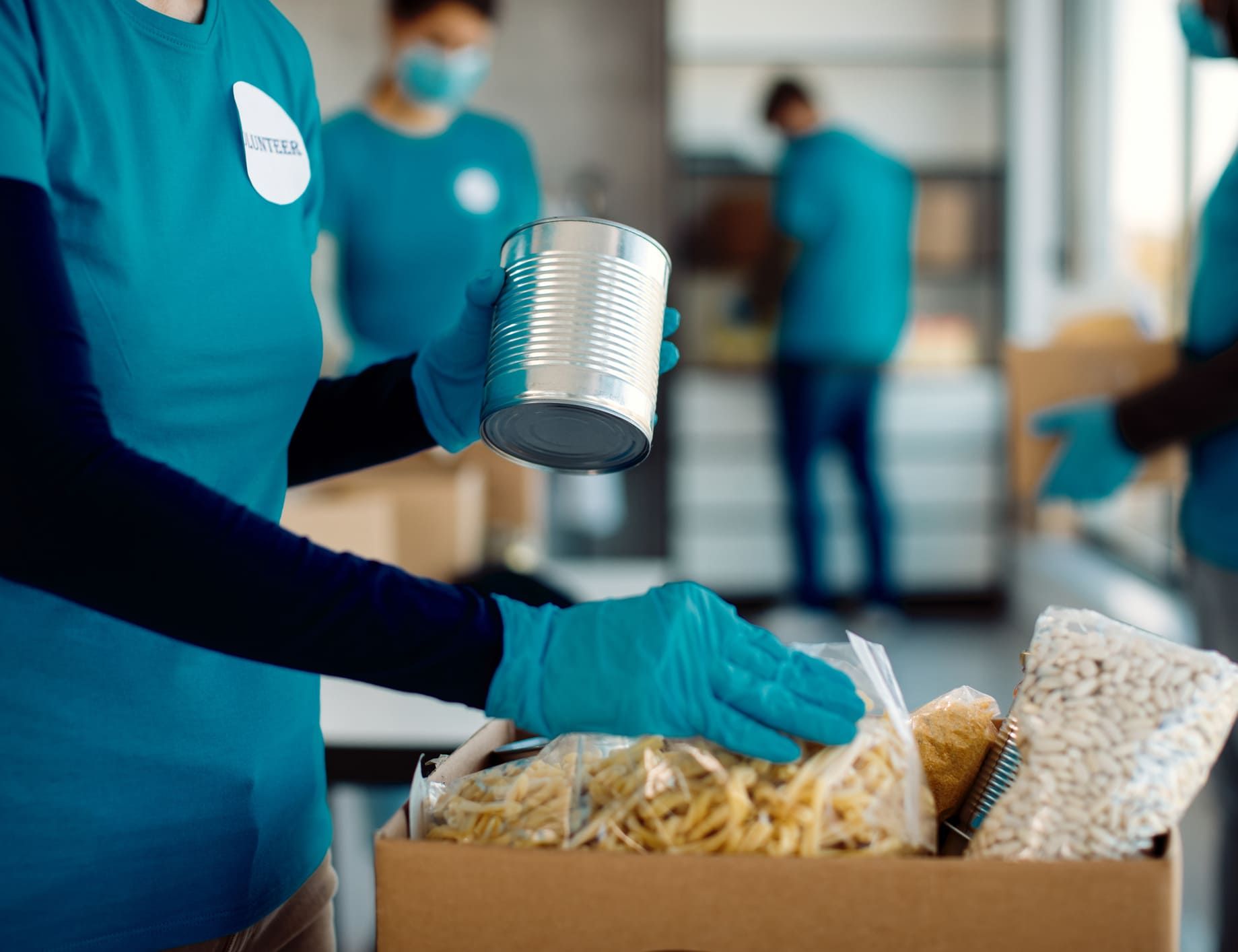Volunteers in teal shirts and gloves pack canned goods and food supplies into a cardboard box for donation.