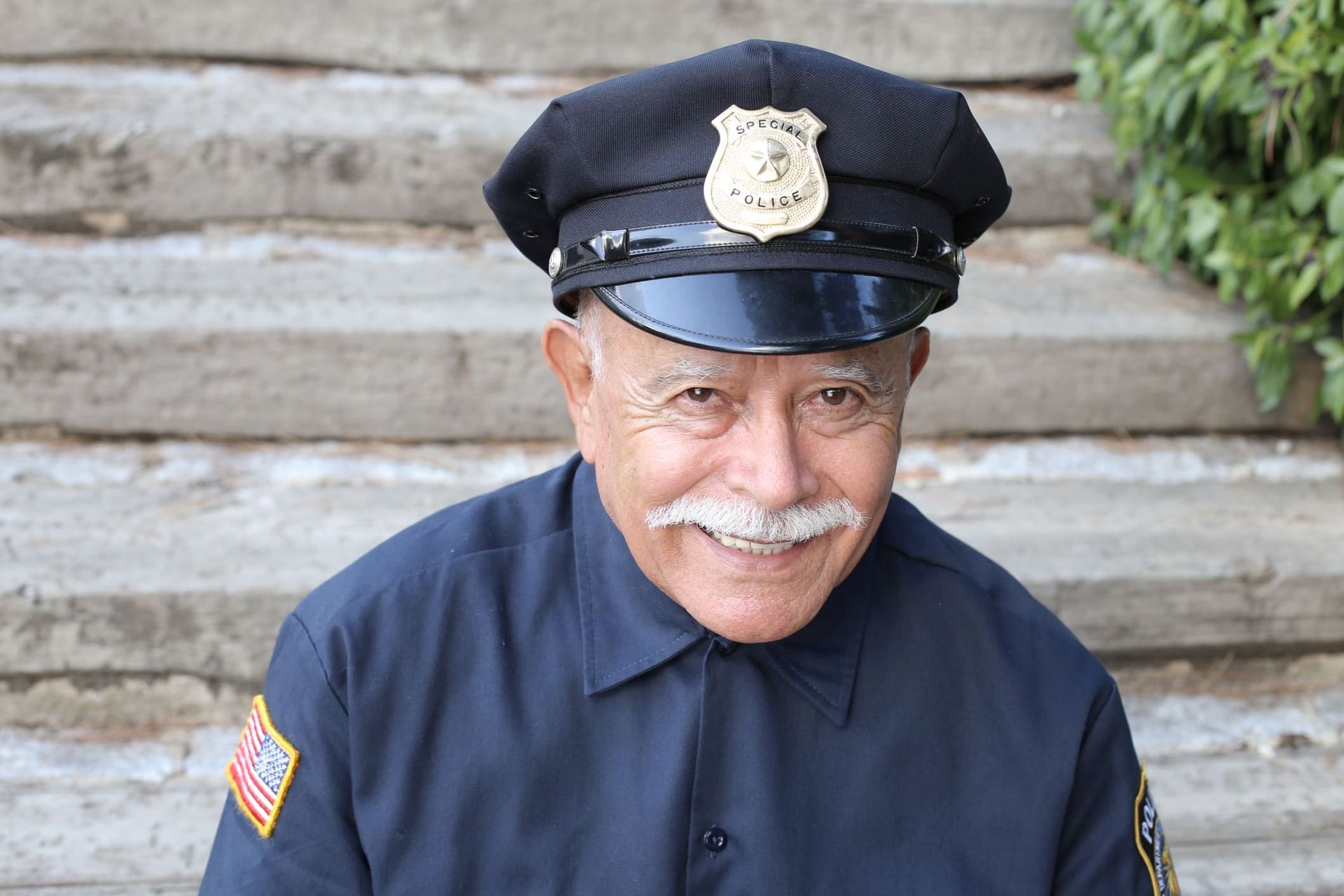 A smiling person in a dark blue uniform and police-style cap sits in front of stone steps.