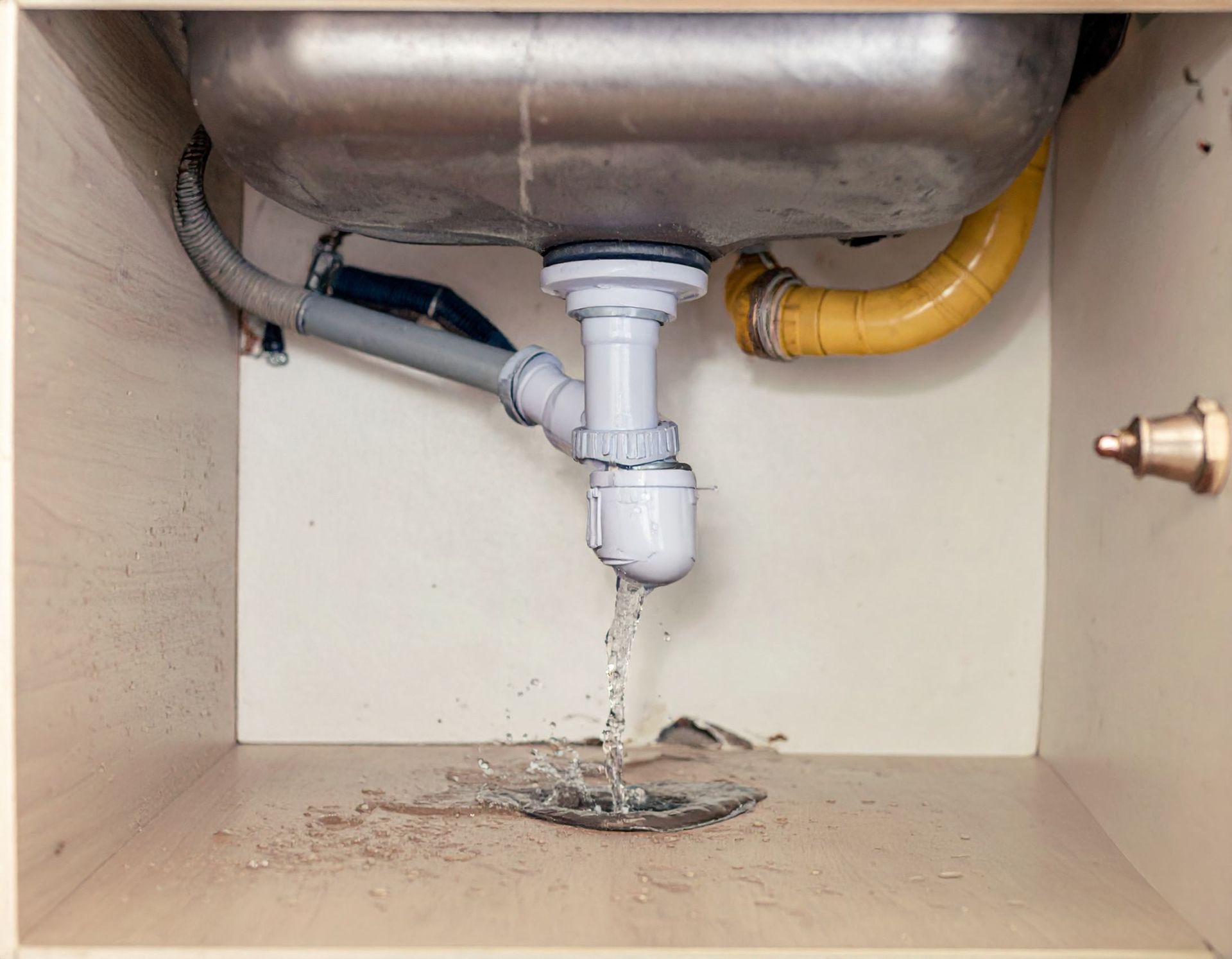 Water leaks from a plastic P-trap pipe under a kitchen sink, pooling on the wooden cabinet floor.