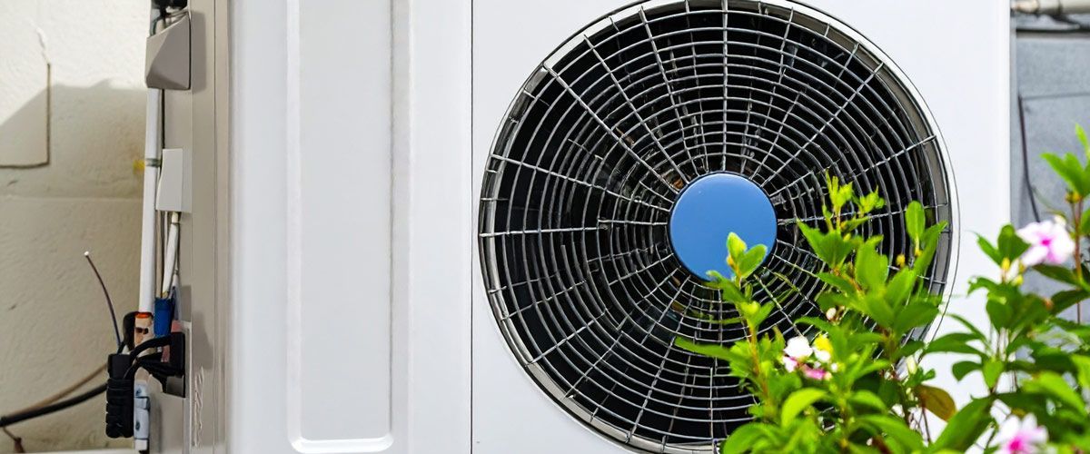 Outdoor HVAC unit with a large black fan beside a white wall and green flowering plant in the foreground