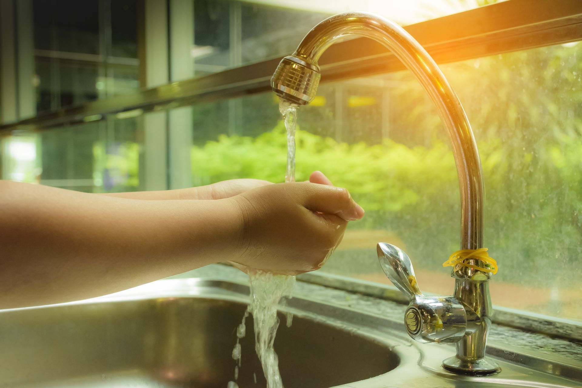 Hands held under a running kitchen faucet, with bright sunlight streaming through a window in the background.