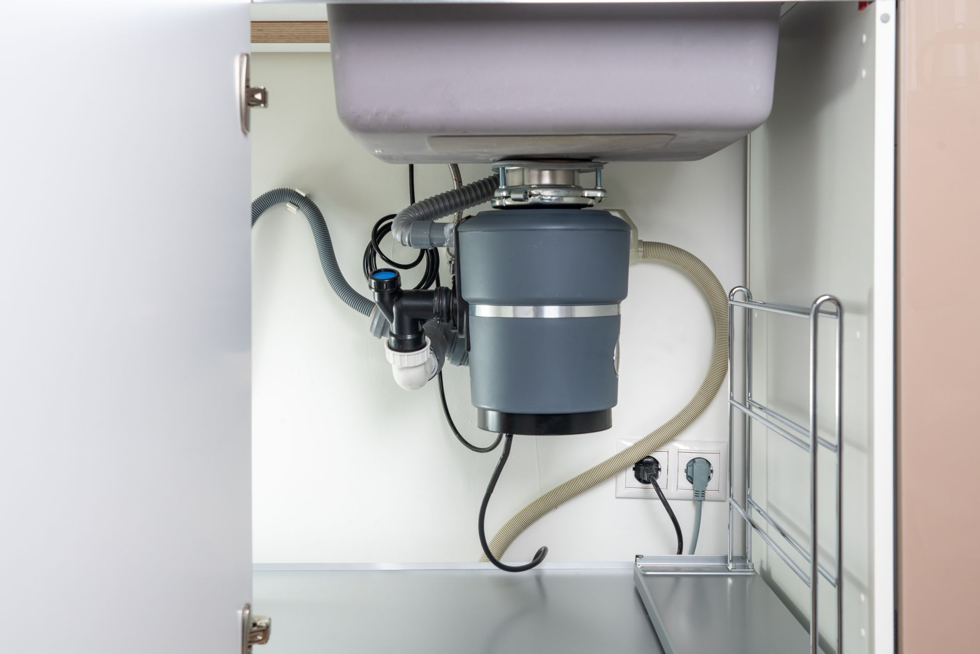 A kitchen sink cabinet interior featuring a gray garbage disposal unit, plumbing connections, and a power outlet.