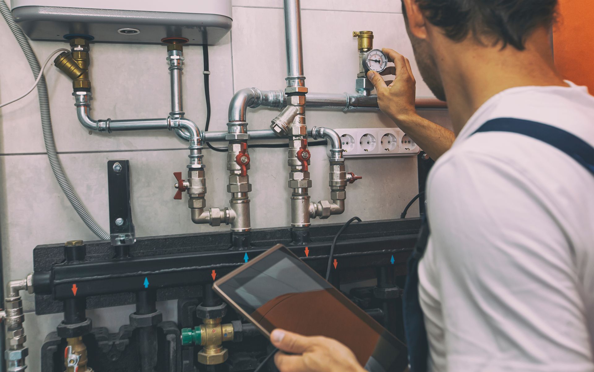 A technician with a tablet checks a wall-mounted heating and plumbing system with pipes, valves, and a water meter.