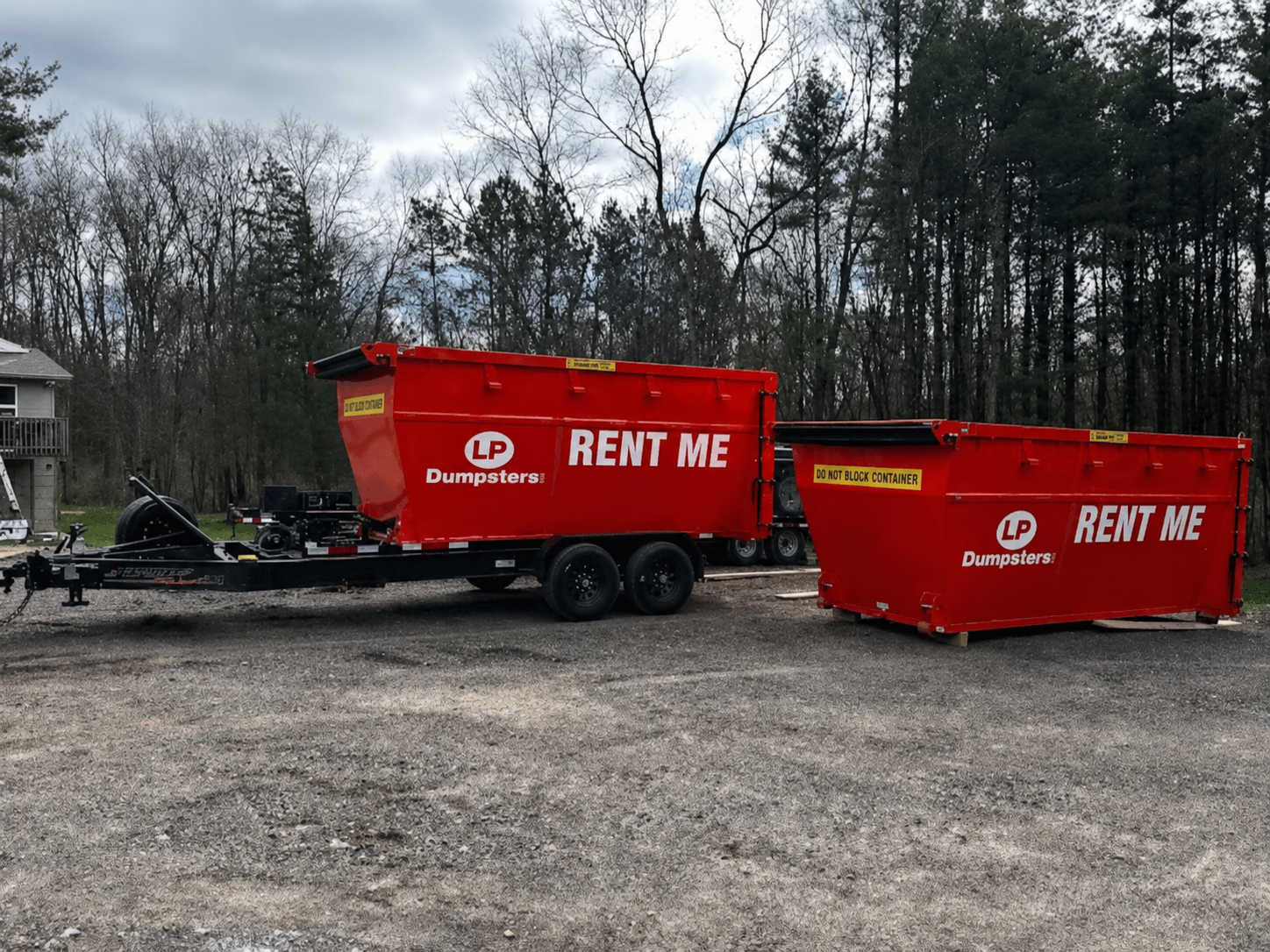 Two red dumpster trailers labeled “RENT ME” parked on a gravel lot by trees.