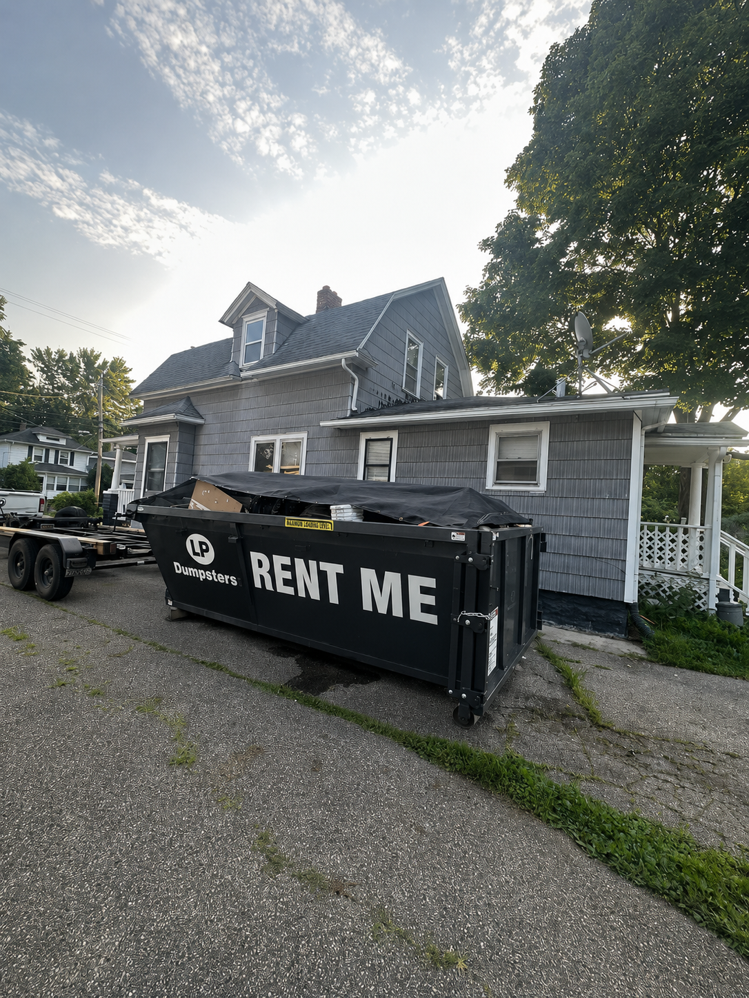 Black dumpster marked “RENT ME” in front of a gray house with a driveway and trees at sunset