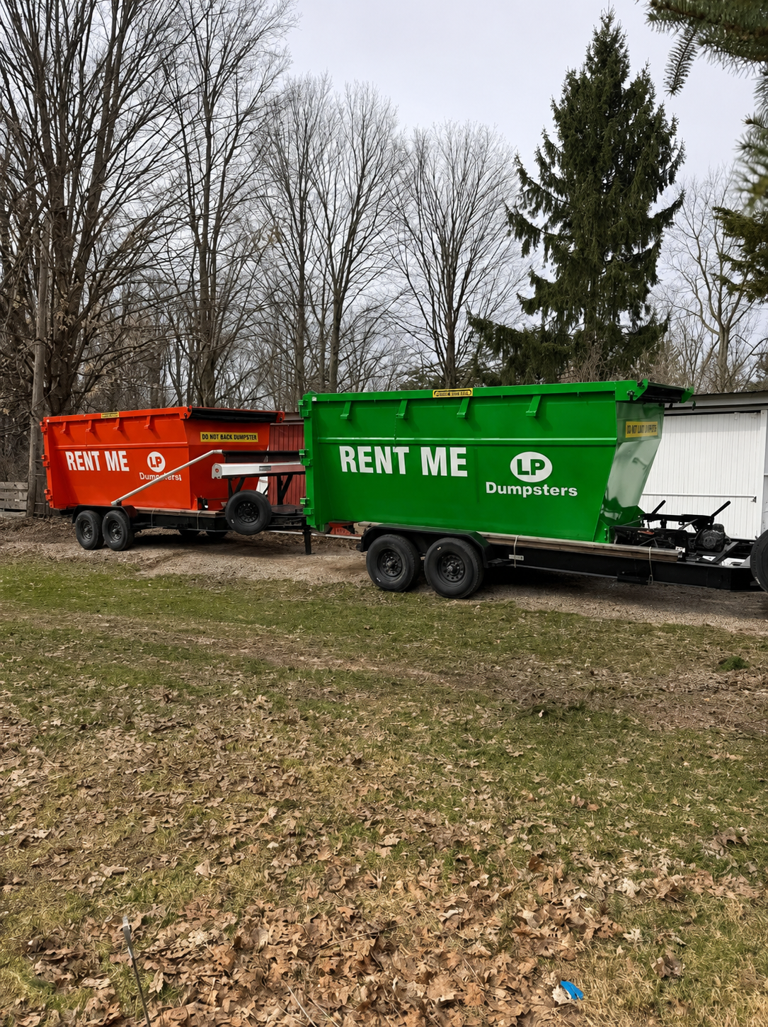 Two dumpsters, one orange and one green, parked on grass by leafless trees.