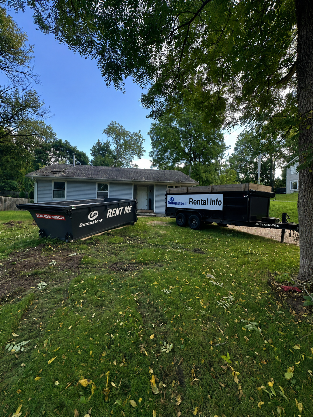 Two black dumpsters beside a house on a grassy yard under trees on a sunny day