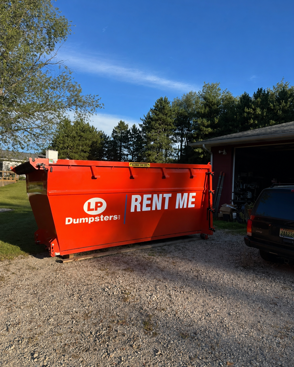 Bright orange dumpster labeled “RENT ME” beside a garage on a gravel driveway.