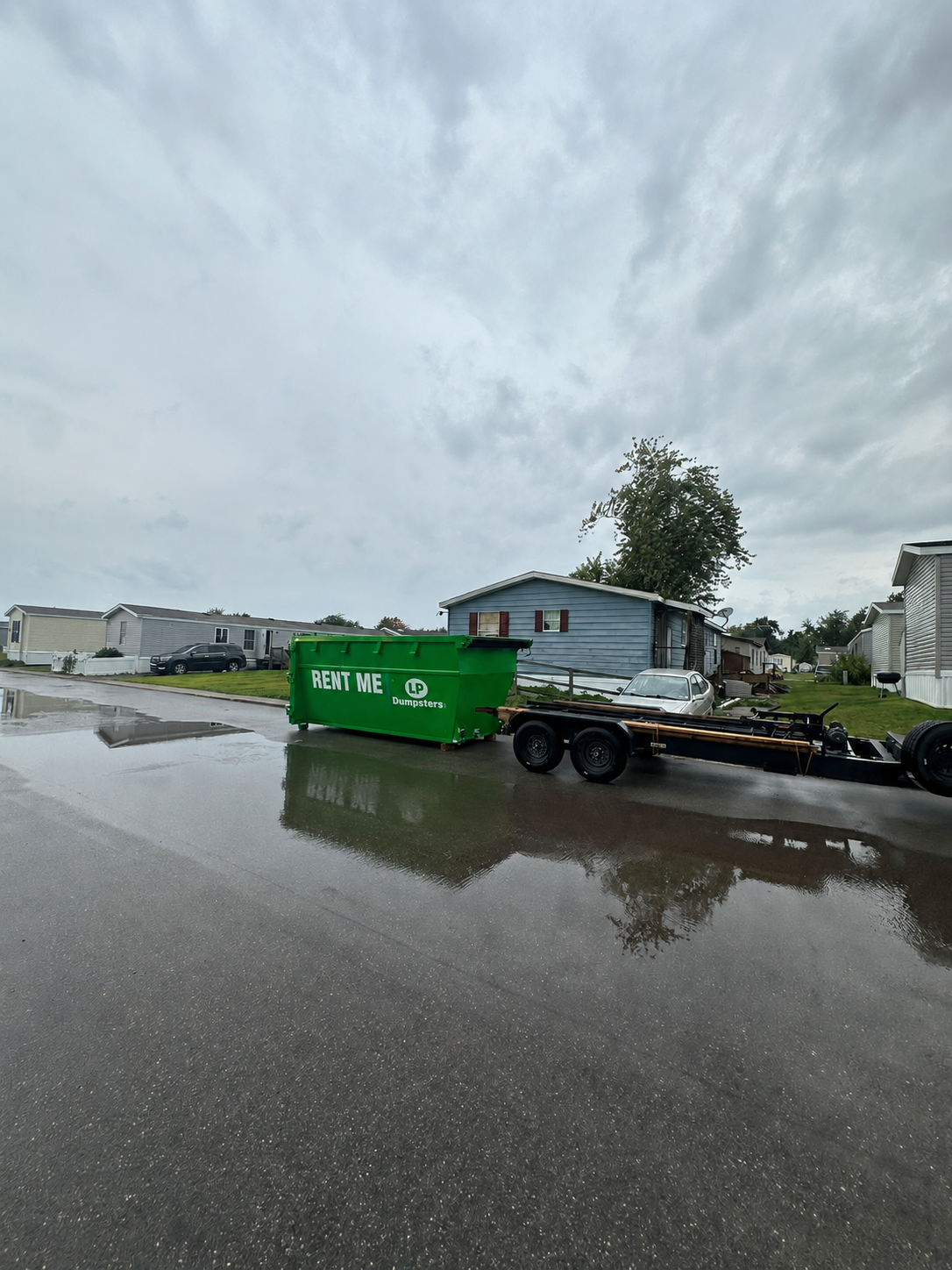 Green dumpster on a trailer in a wet parking lot under cloudy skies, with a house in the background.