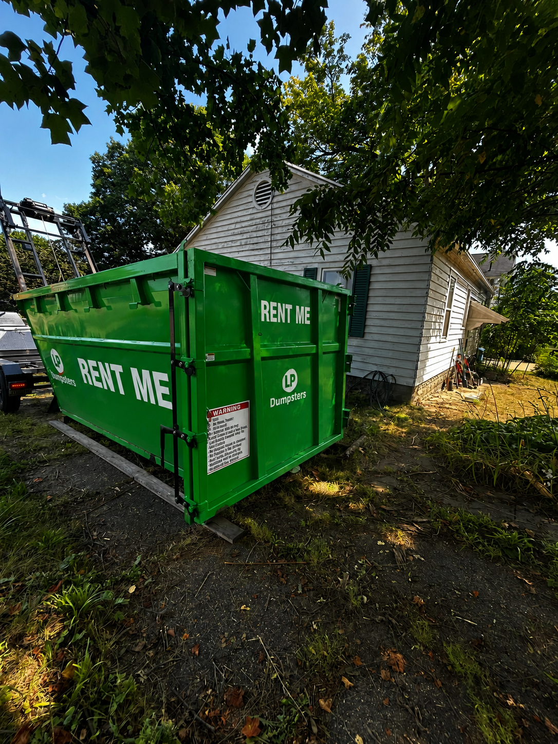 Green FEI container dumpster beside a white house under trees on a driveway.