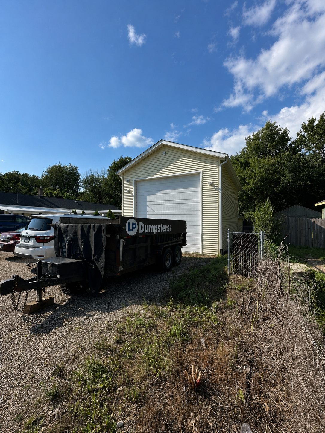 A white shed behind a dumpster in a sunny overgrown yard with trees and a parked car.
