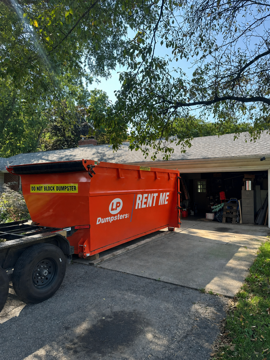Orange rental dumpster beside a garage under trees, with a driveway and open doors in the background