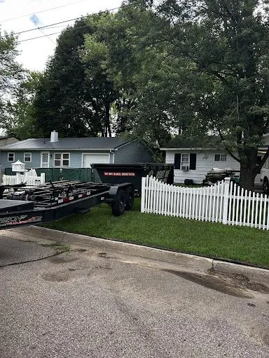 House with large trees, white picket fence, and a black trailer parked by the curb on a residential street.