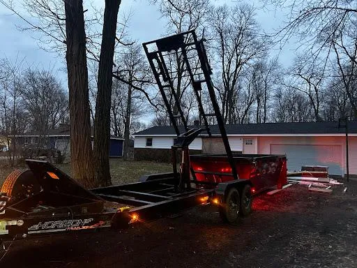 Tow truck with raised wrecker bed and trailer parked at dusk beside a house and leafless trees