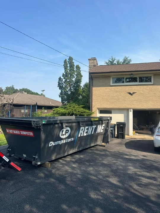 Large black dumpster labeled “RENT ME” parked in a suburban driveway beside a tan two-story house.