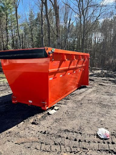 Orange dumpster on dirt lot in a wooded area, with black lid and side panels.