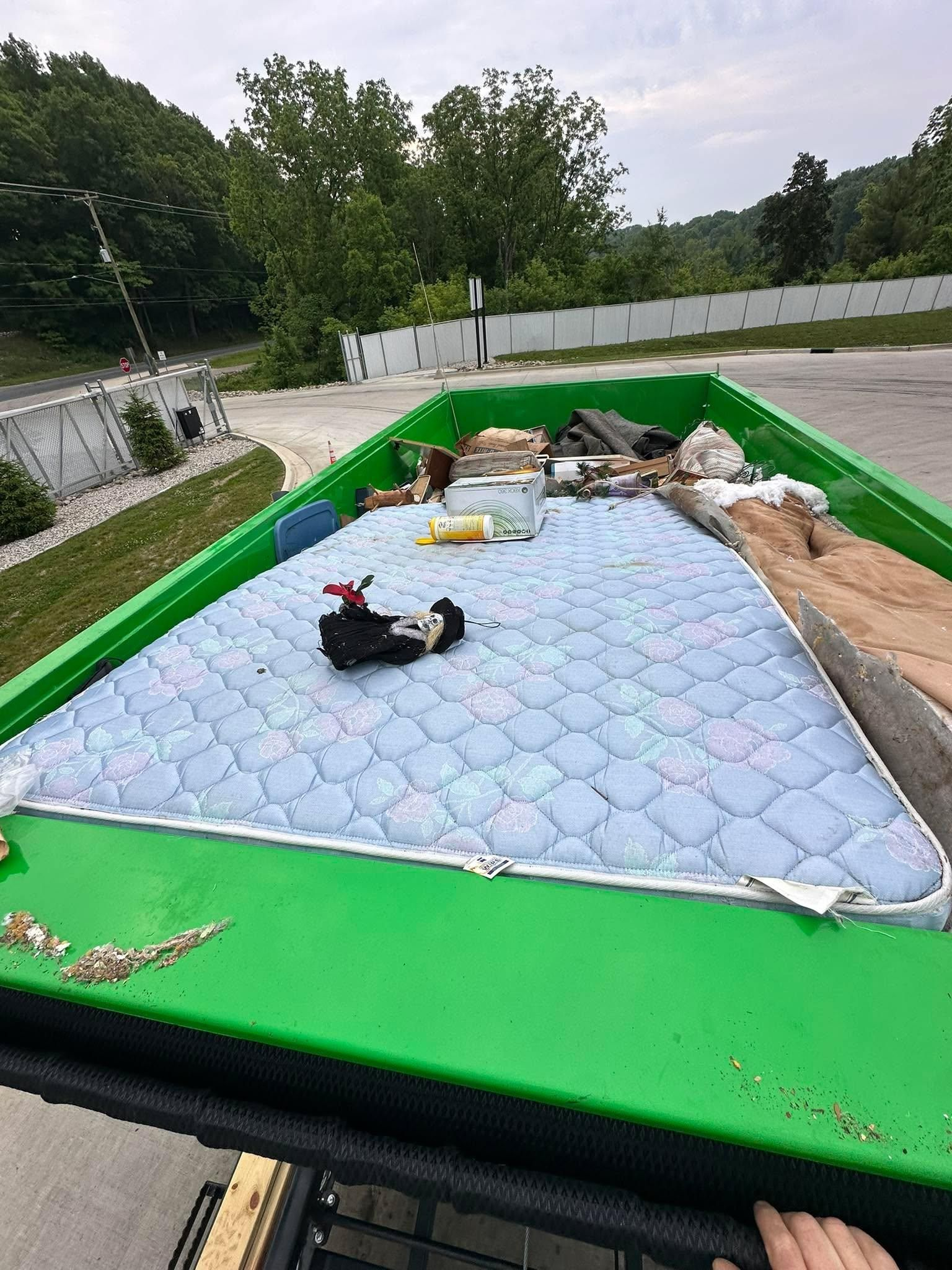 Empty inflatable pool with blue mat, green sides, and scattered items in a wooded outdoor area.