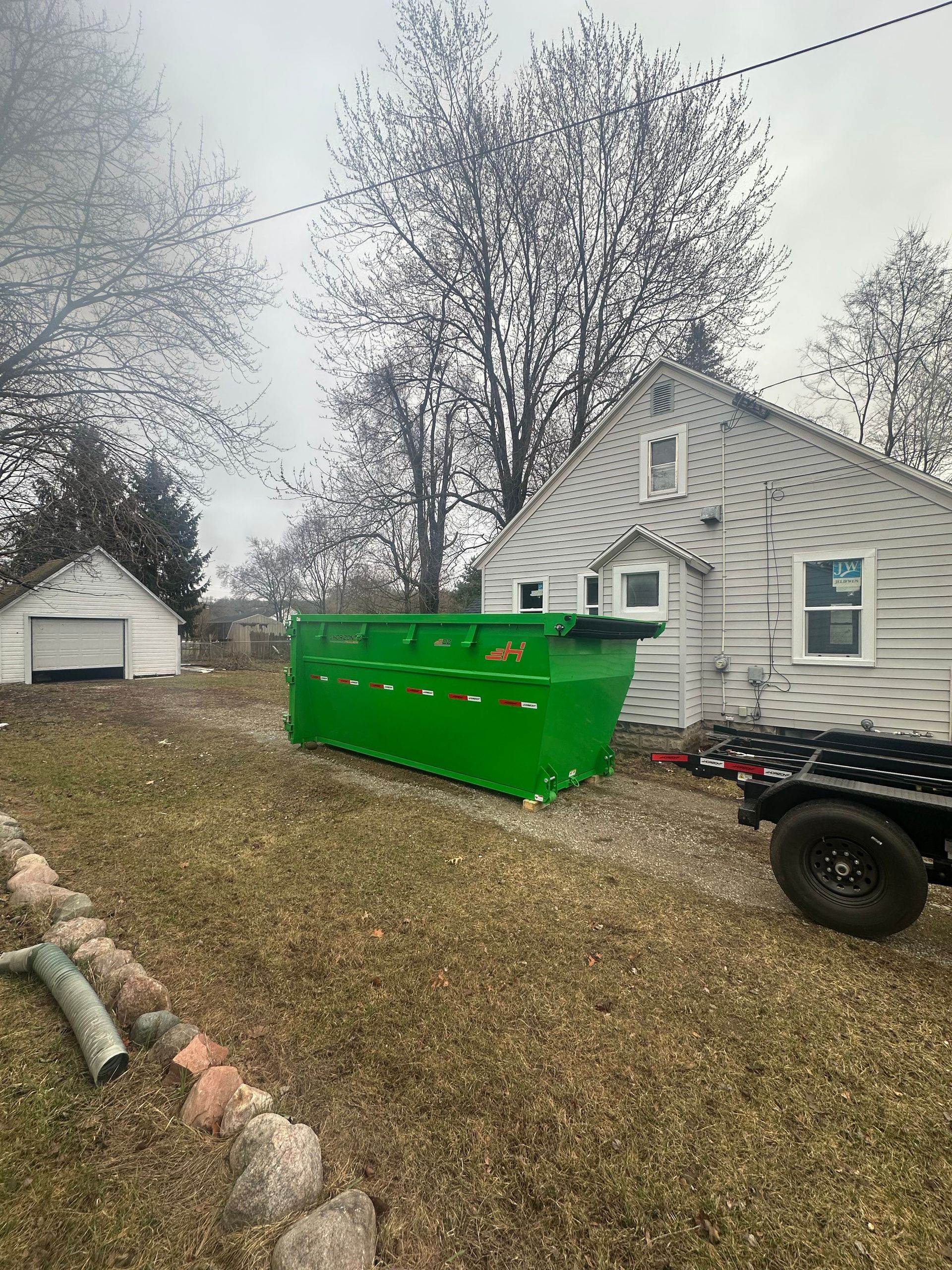 Green dumpster beside a white house and detached garage in a leaf-covered yard