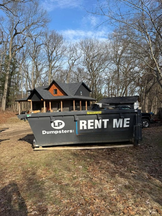 Black dumpster labeled “RENT ME” in front of a house and trees