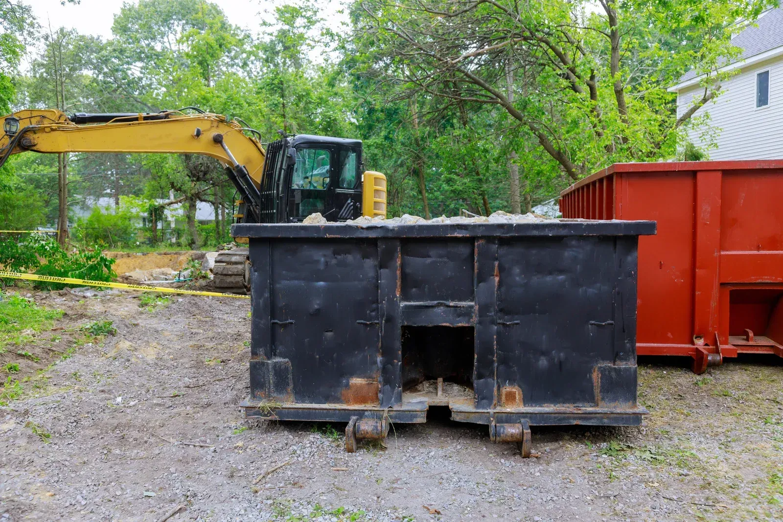 Yellow excavator beside black dumpster and red container in a gravel yard